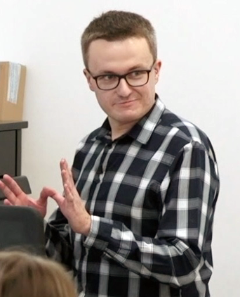 Michał Czubaszek w koszuli w czarno-białą kratę, stojący bokiem przy jasnej ścianie, wykonujący gesty rękami podczas prowadzenia próby.