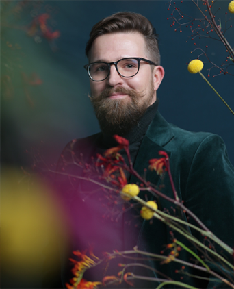 Maciej Leonard Żybula w ciemnozielonej marynarce i czarnym golfie, pozujący na tle ciemnoniebieskiej ściany, otoczony kolorowymi kwiatami o żółtych i czerwonych akcentach..