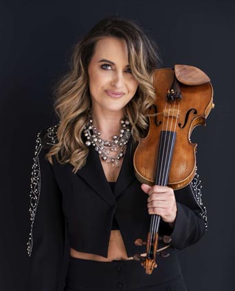 Anna Wandtke, pozująca na ciemnym tle, trzymając skrzypce przy ramieniu. Ma na sobie czarną marynarkę z biżuteryjnymi zdobieniami oraz efektowny naszyjnik.