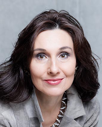 Bliski portret Bogumiły Dziel-Wawrowskiej. Patrzy w obiektyw z delikatnym uśmiechem; ma ciemne włosy i naszyjnik z kryształkami.